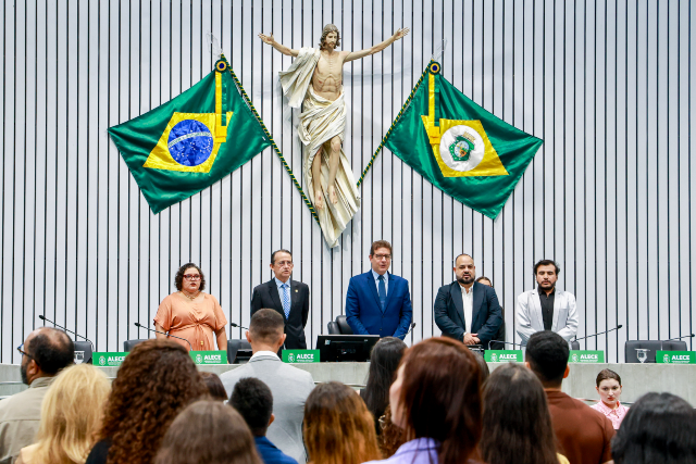 Imagem: No plenário da Assembleia Legislativa do Estado do Ceará (ALECE), seis pessoas estão de pé atrás da mesa principal, voltadas para o público, em postura solene. Ao centro da parede, uma grande escultura de Cristo com os braços abertos está posicionada entre duas bandeiras: a do Brasil e a do Ceará. O público aparece de pé, visto de costas, ocupando a parte inferior da imagem