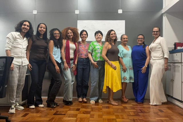 O projeto de extensão O Cuidado como um Direito: Desenvolvimento Econômico e Reconhecimento Jurídico do Trabalho Invisível das Mulheres é uma iniciativa da Escola de Extensão da Faculdade de Direito da UFC (Foto: Divulgação) imagem: Fotografia em ambiente interno. Um grupo de dez pessoas posa lado a lado, sorrindo para a câmera. Há nove mulheres e um homem. Estão em pé sobre piso de madeira, em frente a uma parede cinza com um quadro branco ao centro. As pessoas vestem roupas variadas e coloridas, como calças, blusas, vestidos e sandálias. À direita da imagem, há um móvel branco com objetos apoiados. O clima é descontraído, de registro coletivo.
