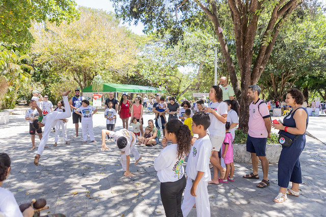 Imagem: Fotografia em espaço aberto e arborizado, durante apresentação de capoeira. No centro da imagem, crianças vestidas com roupas brancas praticam movimentos da capoeira em uma roda improvisada. Ao redor, adultos e crianças acompanham a atividade em pé ou sentados no chão. Algumas pessoas observam atentamente, enquanto outras registram o momento com celulares. O local possui árvores grandes, sombra abundante e clima de convivência comunitária em um dia ensolarado.