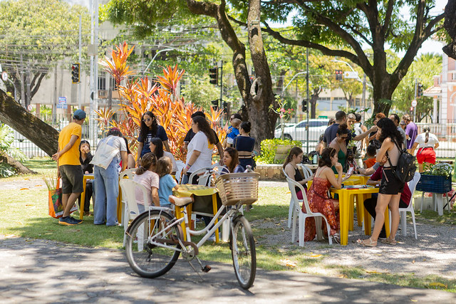 Imagem: Fotografia em área arborizada ao ar livre, durante atividade coletiva. Diversas pessoas, entre jovens e adultos, estão reunidas em mesas e cadeiras espalhadas sobre a grama. Algumas conversam em grupo, enquanto outras participam de atividades manuais. Em primeiro plano, há uma bicicleta parada próxima ao caminho. Ao fundo, árvores grandes fazem sombra sobre o espaço, e é possível ver ruas e construções da cidade ao redor. O ambiente é claro, ensolarado e descontraído.
