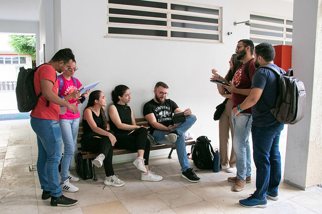 A duração do auxílio é pelo tempo padrão de duração do curso do aluno, definido pela Pró-Reitoria de Graduação (Prograd), prorrogável por até dois semestres (Foto: Ribamar Neto/UFC) Imagem: Fotografia em corredor interno de prédio universitário. Um grupo de oito estudantes conversa e realiza atividades acadêmicas. Três pessoas estão sentadas em um banco, usando cadernos e computador portátil, enquanto as demais permanecem em pé, segurando folhas e conversando entre si. Os estudantes vestem roupas casuais e carregam mochilas. O ambiente possui paredes brancas, piso claro e iluminação natural.