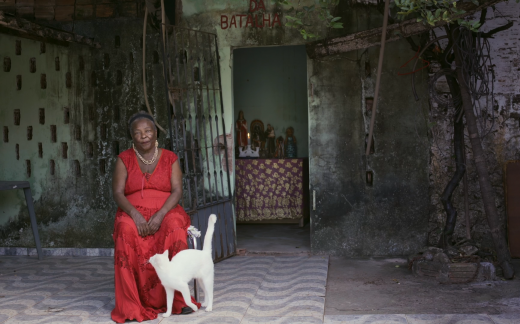 Imagem: imagem do filme nde se vê uma mulher negra, vestida de vermelho, sentada numa cadeira, no que parece ser um quintal de uma casa.