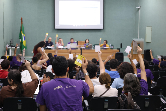 Imagem: Fotografia em auditório durante votação. Diversas pessoas estão sentadas em cadeiras pretas, muitas vestindo camisetas roxas. Vários participantes erguem cartões roxos de votação com o braço estendido. Ao centro da imagem, um homem de barba, também sentado, observa a atividade. Ao fundo, outras pessoas acompanham a sessão. O ambiente é fechado, com paredes claras e iluminação artificial.