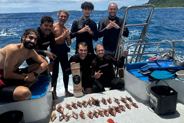 As missões contarão com a participação de pesquisadores e estudantes vinculados ao Labomar e ao Programa de Pós-Graduação em Ciências Marinhas Tropicais (Foto: Arquivo Pessoal) Imagem: Sete mergulhadores posam sobre uma embarcação em alto-mar, sob céu parcialmente nublado. O grupo, formado por homens, veste roupas de mergulho pretas; alguns estão descalços e outros usam parte do equipamento. Todos sorriem para a câmera e fazem gestos com as mãos. À frente deles, sobre o piso branco do barco, estão dispostos vários peixes-leão alinhados em fileiras, resultado da ação de manejo da espécie invasora. Também aparecem equipamentos de mergulho, como nadadeiras azuis, cilindro e baldes. Ao fundo, o mar azul se estende até uma faixa de vegetação em terra firme, visível no lado direito da imagem.