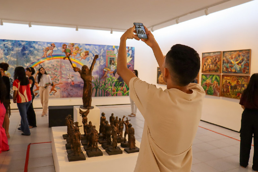 Imagem: A imagem mostra o interior de uma galeria do Museu de Arte da UFC, com visitantes observando uma exposição de pinturas e esculturas. Em primeiro plano, aparece um homem de costas, usando camisa clara, segurando um celular acima da cabeça para fotografar ou filmar as obras expostas. À frente dele, sobre uma base branca, há diversas esculturas pequenas em tom escuro, organizadas em conjunto, além de uma escultura vertical maior ao centro. Nas paredes ao fundo, destacam-se pinturas coloridas e expressivas, com cenas figurativas e elementos humanos. À esquerda, um grande quadro em tons de azul, lilás e rosa ocupa boa parte da parede. À direita, há uma sequência de telas menores com cores quentes e composição vibrante. Outros visitantes circulam pelo espaço, observando as obras e conversando. O ambiente é amplo, iluminado por luzes direcionadas no teto, com paredes brancas e atmosfera contemplativa típica de um museu de arte contemporânea.
