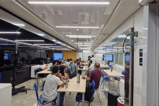 As atividades estão acontecendo dentro do caminhão-laboratório do projeto, estacionado no bloco 942-A do Campus do Pici (Foto: Divulgação) Imagem: imagem de dentro de uma sala de aula com mesas, cadeiras, estudantes de costas olhando pra uma lousa e sentados ao redor de mesas. Local com janelas de vidros.