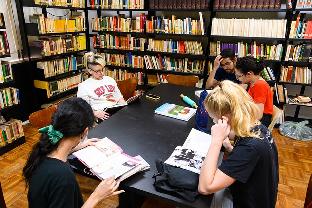 Imagem: Sala de biblioteca com estantes altas repletas de livros organizados ao fundo e nas laterais. No centro, uma mesa retangular preta reúne cinco estudantes sentados, concentrados na leitura e consulta de materiais.  Uma estudante de cabelos escuros presos por um laço verde folheia um livro aberto. À frente, outra estudante loira consulta um livro, apoiando uma das mãos no rosto. Ao lado, dois estudantes observam o mesmo material, enquanto uma estudante de cabelos claros, sentada ao fundo, lê um livro com atenção. Sobre a mesa, há livros, cadernos, estojo e garrafas. O ambiente é iluminado, silencioso e organizado, sugerindo momento de estudo coletivo e pesquisa acadêmica.