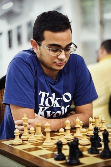 Imagem: Jovem enxadrista participa de uma partida de xadrez em ambiente interno. Ele tem cabelos curtos escuros, usa óculos e veste camiseta azul com a inscrição “Tokyo Kyoto”. Sentado diante do tabuleiro, mantém os braços cruzados sobre a mesa e observa atentamente as peças, demonstrando concentração.  Em primeiro plano, aparecem peças brancas e pretas distribuídas no tabuleiro durante a partida. Ao fundo, o ambiente está desfocado, com outras pessoas sentadas, sugerindo um torneio ou espaço coletivo de jogos.