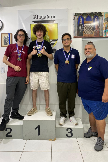 Imagem: Quatro homens posam em uma cerimônia de premiação de xadrez. Três deles estão sobre um pódio branco numerado, usando medalhas no peito. O primeiro colocado, ao centro, está na posição mais alta do pódio, veste camiseta preta e bermuda clara, e segura a medalha com as duas mãos. À esquerda, o segundo colocado usa camiseta vinho e calça escura. À direita, o terceiro colocado veste camisa azul-marinho e calça verde-escura.  Ao lado do pódio, um homem de barba grisalha, usando camisa polo azul e bermuda azul, posa sorrindo para a foto. Ao fundo, há quadros decorativos relacionados ao xadrez e um banner com a palavra “Alagadiço”. O ambiente é interno, com piso claro e iluminação branca.