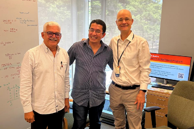 Aldo Ângelo Moreira Lima, Francisco de Sousa Junior e Eric R. Houpt na University of Virginia, Charlottesville, EUA, em 20 de maio de 2026 (Foto: Divulgação) Imagem: Três homens adultos posam sorrindo em um escritório iluminado. À esquerda, um homem de cabelos brancos e óculos veste camisa social clara. No centro, um homem de camisa azul listrada abraça os colegas pelos ombros. À direita, um homem careca usa camisa branca social e crachá pendurado no pescoço. Ao fundo, há uma grande janela com vista para árvores, uma mesa de escritório com computador e cadeira, além de um quadro branco com anotações e fórmulas escritas em diferentes cores. Na tela do computador, aparece uma apresentação relacionada a saúde e bem-estar. O ambiente transmite contexto acadêmico, científico ou institucional.