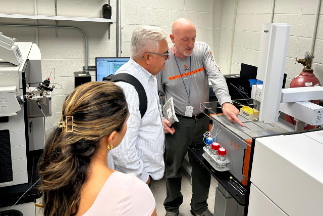 Aldo Ângelo e Nicholas E. Sherman, Associate Professor of Microbiology e Core Director do Biomolecular Analysis Facility Core (BAF) da University of Virginia (Foto: Divulgação) Imagem: Laboratório com equipamentos científicos e computadores. Em primeiro plano, uma mulher de cabelos presos observa dois homens ao lado de um equipamento de análise laboratorial. Um dos homens, de cabelos brancos e óculos, segura documentos enquanto acompanha a explicação do outro pesquisador, um homem careca que aponta para a máquina. O equipamento possui compartimentos transparentes, frascos com líquidos e estruturas metálicas, sugerindo atividade de pesquisa ou demonstração técnica. Ao fundo, aparecem computadores, outros aparelhos laboratoriais e cilindros de gás, compondo um ambiente científico e tecnológico. Os participantes usam roupas casuais e crachás, indicando contexto acadêmico ou institucional.