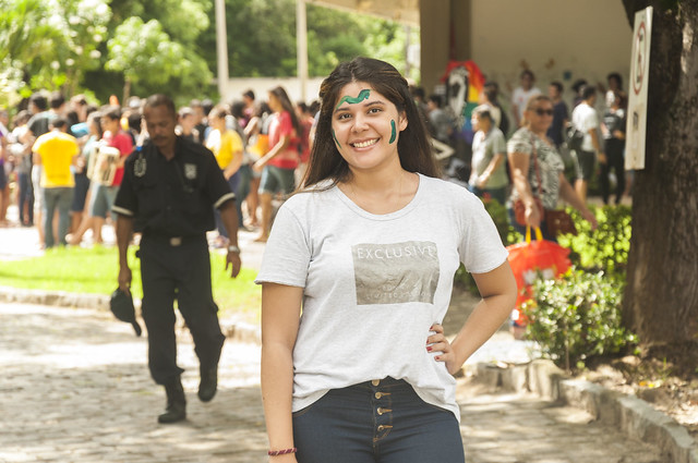 Imagem: mulher jovem sorrindo para a câmera em um ambiente externo com várias pessoas ao fundo. Ela usa camiseta cinza e calça escura, está com a mão na cintura e possui pinturas verdes no rosto. Ao fundo, há árvores, pessoas caminhando e um agente de segurança passando pelo local