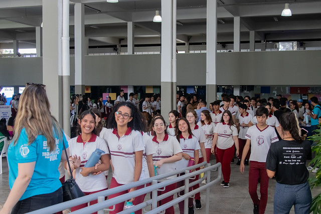 imagem: Espaço amplo e coberto de uma instituição de ensino, com grande circulação de estudantes. Em primeiro plano, um grupo de jovens uniformizados caminha por uma rampa, sorrindo e conversando. Eles vestem camisetas brancas com detalhes em vinho e calças da mesma cor, típicas de uniforme escolar.  À esquerda, uma mulher de costas, usando camiseta azul, parece orientar ou recepcionar os participantes. Ao fundo, dezenas de estudantes ocupam o ambiente, formando filas e grupos em circulação. O local possui pilares altos, iluminação suspensa e áreas abertas, criando um clima de evento educacional movimentado.  A cena transmite um momento de integração e participação estudantil, possivelmente relacionado a uma feira, mostra de profissões ou atividade de acolhimento acadêmico.