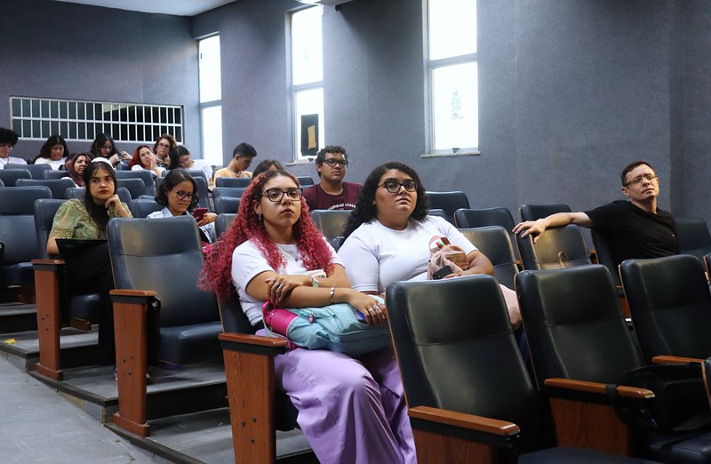 Imagem: Auditório com paredes na cor cinza, com público do seminário sentado em cadeiras acolchoadas de cor preta e com pés de madeira.