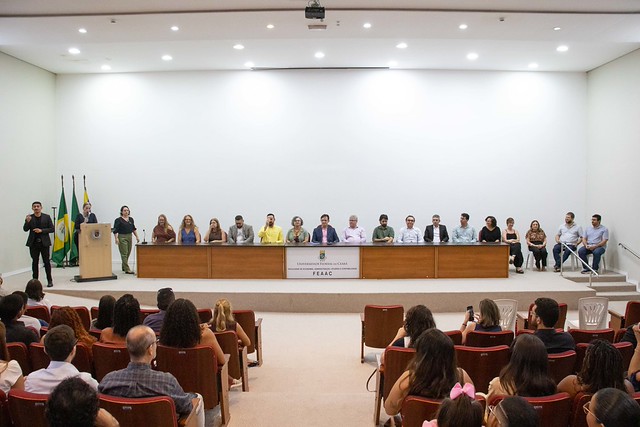 Imagem: auditório durante uma cerimônia acadêmica. No palco, diversas pessoas estão sentadas atrás de uma longa mesa, compondo a mesa de honra do evento. À esquerda, uma pessoa fala ao microfone em um púlpito. Na plateia, participantes acompanham a solenidade