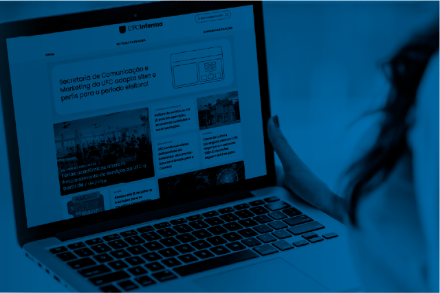 Imagem: Fotografia em tons de azul mostra uma pessoa utilizando um notebook sobre o colo. Na tela está aberta a página inicial do portal UFC Informa, com destaques de notícias organizados em blocos. A mão direita da pessoa segura a lateral do computador, enquanto parte do rosto e dos cabelos aparece desfocada à direita da imagem. O teclado do notebook ocupa a metade inferior da fotografia, e o fundo está desfocado, dando destaque ao equipamento e ao portal exibido na tela.