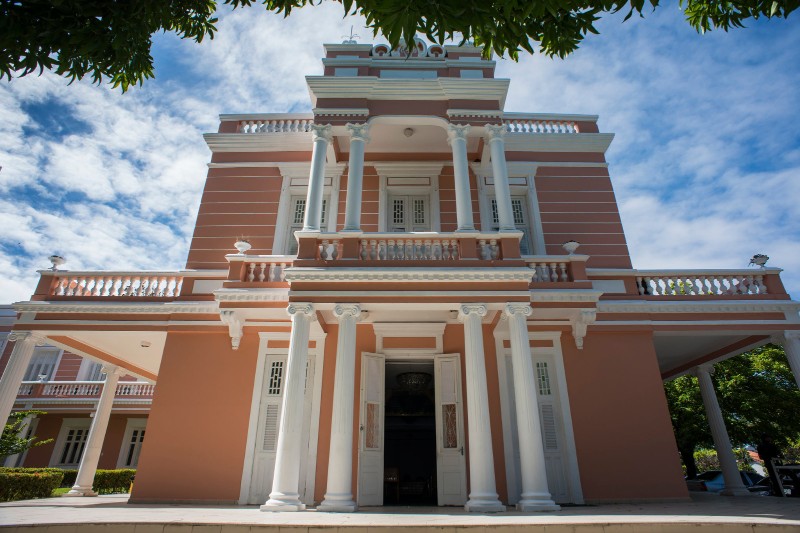 Imagem: fachada da reitoria que abriga o auditório, um prédio com dois pavimentos na cor rosa com detalhes em branco.