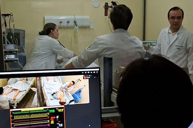 Imagem: Fotografia mostra uma atividade de simulação clínica em ambiente hospitalar. Em primeiro plano, vê-se a sala de controle da simulação, com monitores de computador ligados e a silhueta de uma pessoa observando a cena. Um dos monitores exibe a imagem de um manequim de simulação deitado em um leito hospitalar. Ao fundo, separados por um vidro, quatro profissionais de saúde usam jalecos brancos e participam da atividade em uma sala de atendimento. Um deles gesticula enquanto conversa com a equipe; outro está de costas para a câmera; uma profissional atende o paciente simulado junto ao leito; e outra aparece parcialmente à direita da imagem. Equipamentos médicos e um monitor de sinais vitais completam o cenário, característico de um treinamento em ambiente de terapia intensiva.