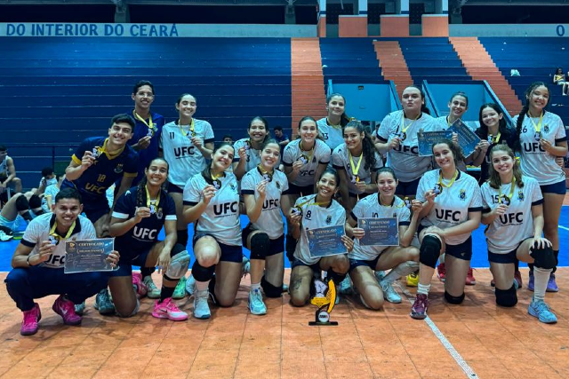 Imagem: Trata-se de uma foto das atletas e da equipe técnica da seleção feminina de vôlei da UFC reunidas em quadra, exibindo medalhas, troféu e certificados conquistados. O grupo posa sorridente em um ginásio, celebrando o resultado da competição.