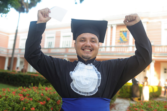 Imagem: jovem pardo sorridente veste beca preta com faixa azul, com as mãos para o alto. Ao fundo aparece a fachada rosa da Reitoria da UFC
