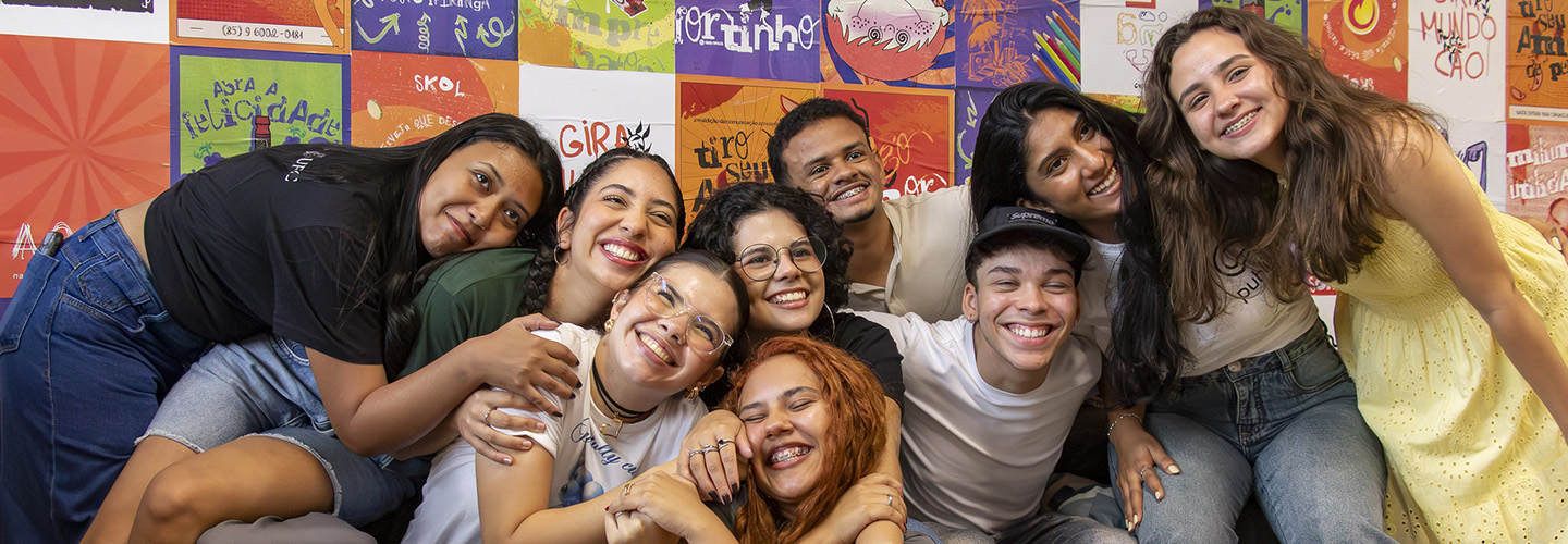 Grupo de nove alunos se abraçam e sorriem tendo ao fundo um painel multicolorido