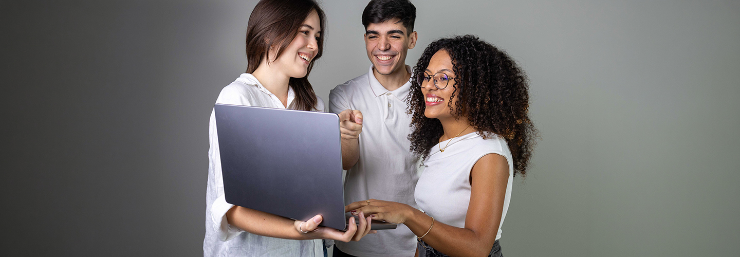 Três estudantes vestindo roupas brancas se olham e sorriem, e segurado um laptop em estúdio fotográfico