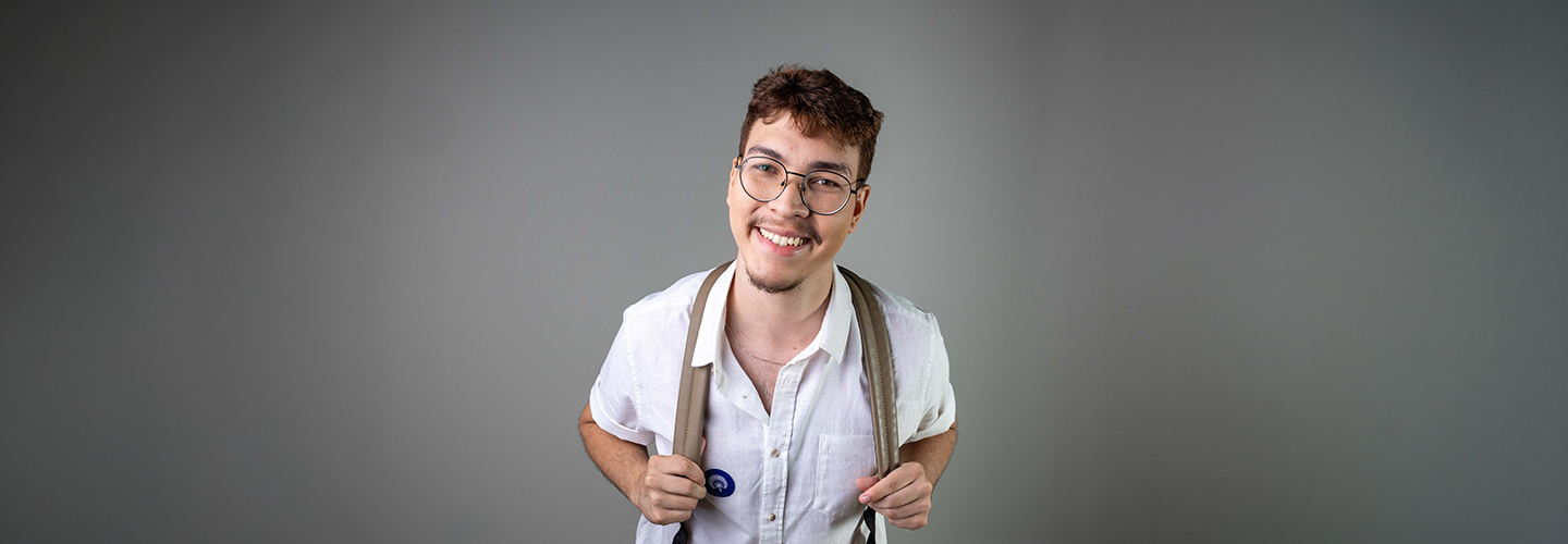 Aluno branco usando óculos e mochila, vestindo camisa branca, posa em estúdio fotográfico