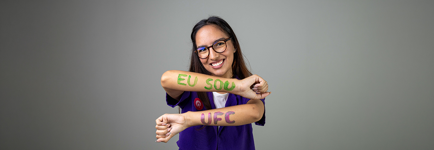 Aluna negra com óculos posa em estúdio fotográfico; em seus braços há pintada com tinta a frase "Eu sou UFC"