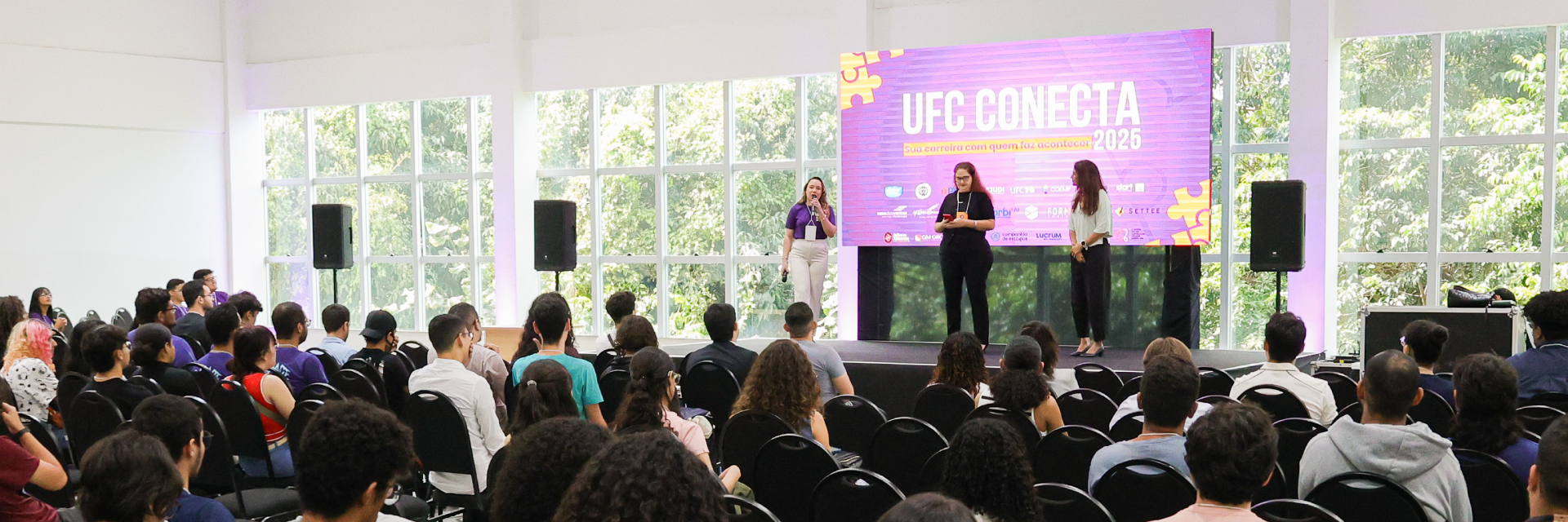 Foto de auditório com público sentado diante de um palco, onde três mulheres estão em pé. Ao fundo, um telão exibe a identidade visual do UFC Conecta 2026