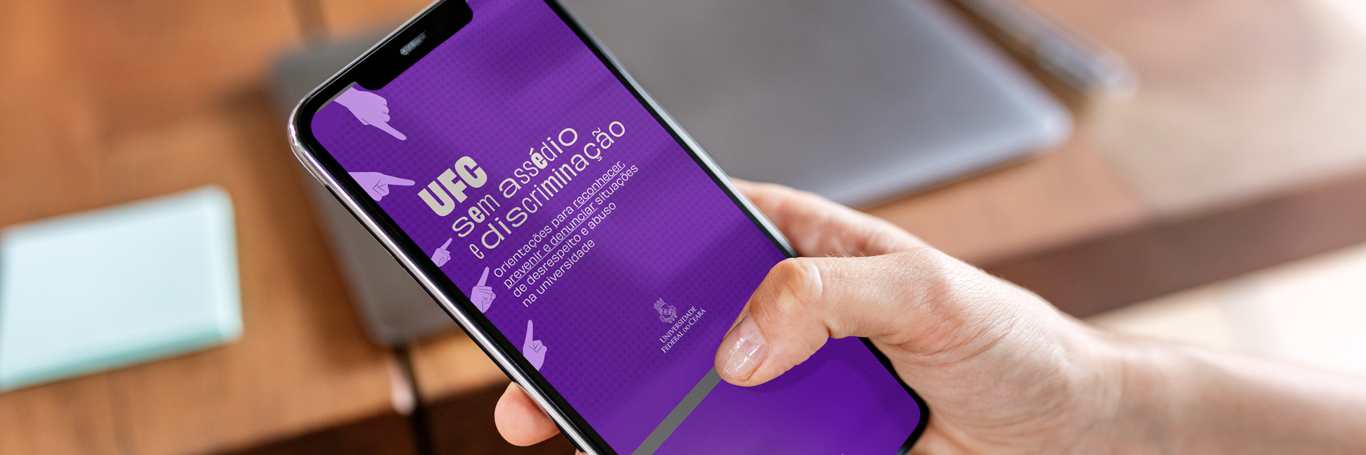 Card com a mão de uma pessoa segurando um celular; na tela, a capa em fundo roxo do guia UFC Sem Assédio e Discriminação